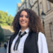 A young Caucasian woman with curly brown hair, smiling slightly off camera. She is wearing a white shirt, black ribbon tie, and black academic gown, standing outdoors with blurred college buildings and trees in the background