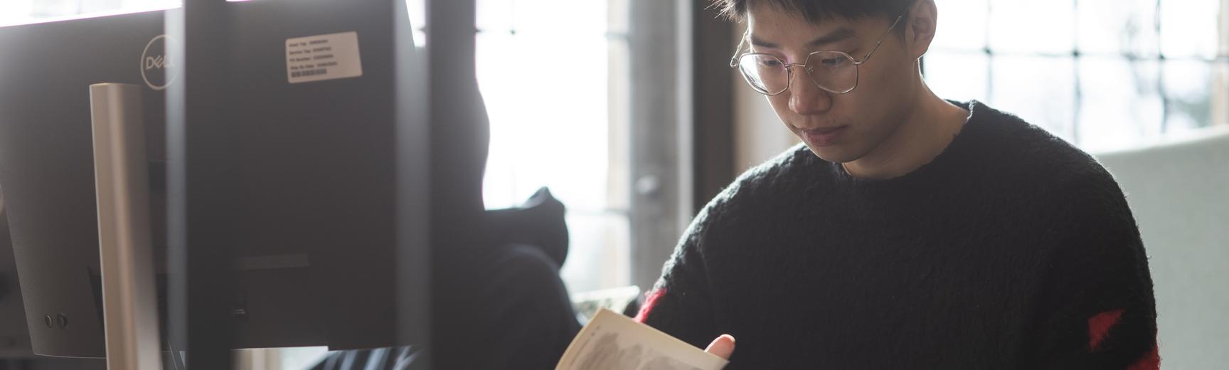 A student wearing glasses and a dark jumper sits at a desk in a bright room, reading a book. A computer monitor is visible to the left, and large windows let in natural light in the background.