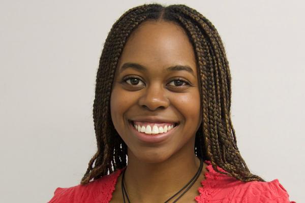 A headshot of a young Black woman with brown braids smiling. She is wearing a bright red top and round necklace.