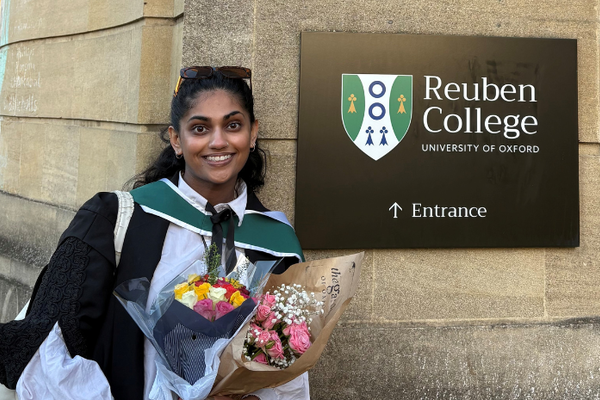Shreya Ganguli, a woman of South Asian descent with long dark hair, smiles while holding a bouquet of flowers in front of a stone wall featuring the Reuben College sign. She is in a graduation gown.