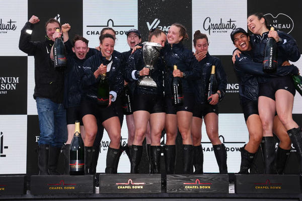 Oxford Women's Blue Boat crew celebrating on the podium with trophy and champagne, smiling and cheering together.