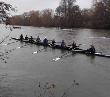 A crew of rowers rowing out on the water. The sky is grey and they're wearing hoodies.