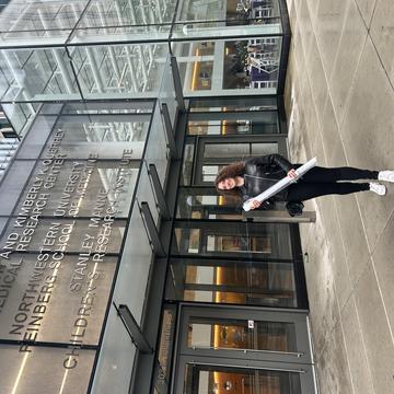 A woman standing outside the Louis A. Simpson and Kimberly K. Querrey Biomedical Research Center at Northwestern University, smiling and holding a rolled-up poster on a rainy day.