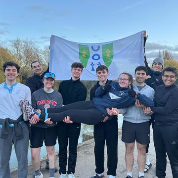 A group of graduate students by the river posing together. One at the back lifts a flag with the Reuben crest