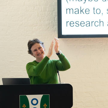Vice-Chancellor, Prof Irene Tracey, stands at a lectern bearing the Reuben College crest, smiling and applauding during her Tuesday Talks lecture.