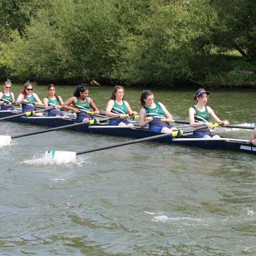 The Reuben women’s rowing crew mid-race on the river, all in matching green vests and blue shorts, rowing in sync. Among the team is Shreya.