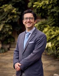 A Caucasian man with short, dark hair swept back. He's dressed professionally in glasses, a blue shirt and tie, and a grey suit jacket. 
