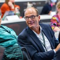 Andrew Thompson, a smartly-dressed Caucasian man with short hair and glasses. He is smiling and turning away from his microphone.