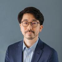 A man of Asian descent with dark hair, glasses, and a stubbled beard. He is wearing glasses and a navy suit. 