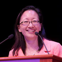Dr Vivian Lee, a smartly-dressed woman of Asian descent with shoulder-length black hair. She is speaking at a podium and smiling.