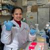 A young woman in the lab, squeezing a pipette into a test tube. She has light brown skin and curly black hair, and is wearing blue gloves and a white lab jacket. She is smiling at the camera.