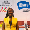A young Kenyan woman in a yellow top smiles confidently at the AFS Youth Assembly. Behind her are festive banners and international flags.