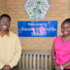 Two people smiling outside Stanton Harcourt School's sign, standing by a wooden bench. The person on the left is wearing glasses, a yellow sweater, and dark trousers. The right person wearing a bright pink top and dark trousers.