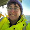 A Caucasian woman with glasses and short brunette hair smiling. She is in a fluorescent yellow jacket and is sitting on a boat with the sea behind her. 